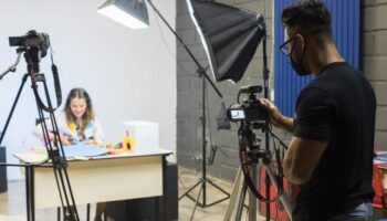 Imagem mostra cinegrafista de máscara com uma camêra e uma mulher em uma mesa sendo gravada. O cenário de como gravar aulas on-line.