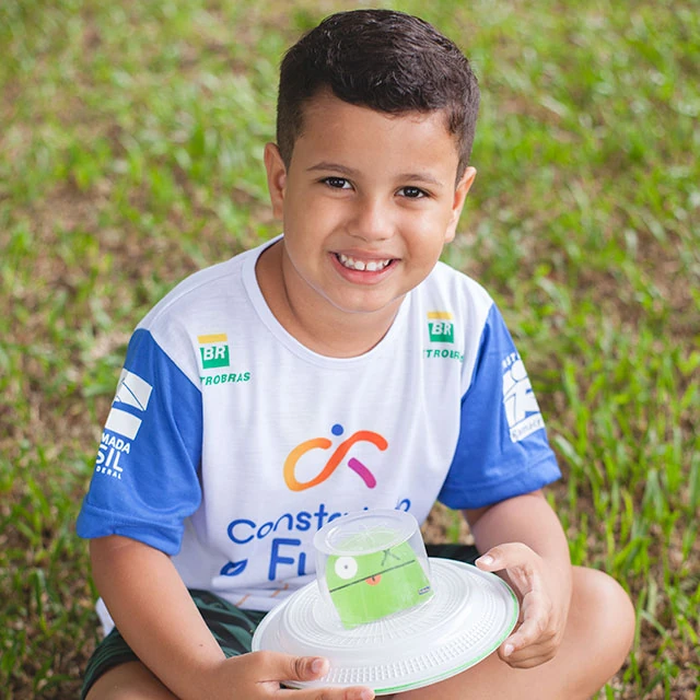 Menino sentado na grama olhando para a câmera e sorrindo, com uniforme do Projeto Construindo o Futuro patrocinado pela Petrobras, segurando brinquedo feito a partir de material reaproveitado