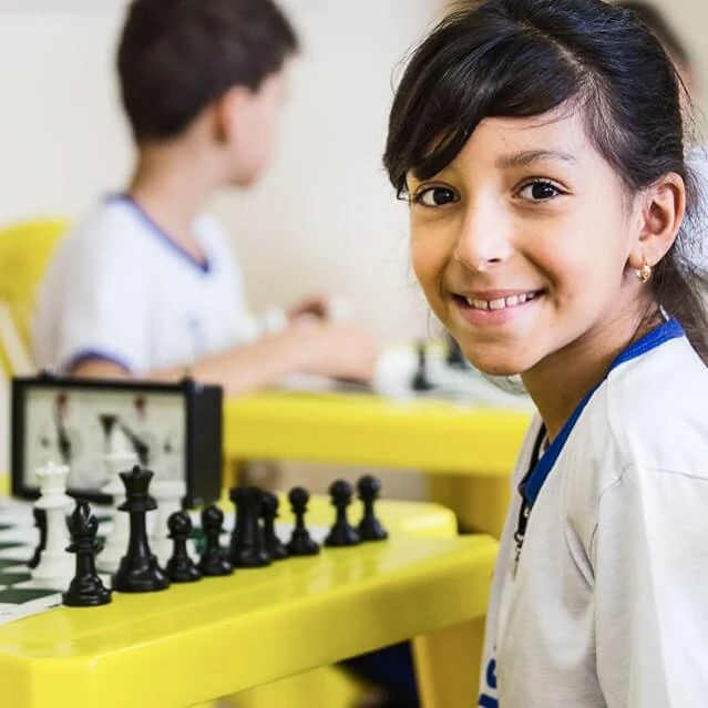 Aluna do Instituto Ramacrisna, olhando de lado para a câmera e sorrindo, com uma mesa de xadrez à frente, ao fundo, outro aluno numa mesa de xadrez